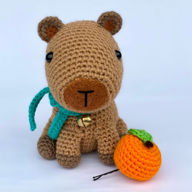 Crochet sculpture of a sitting capybara with an oversize head and smaller body. Next to it is an orange citrus fruit attached to a bobby pin and around its neck is a teal scarf and a metal bell.