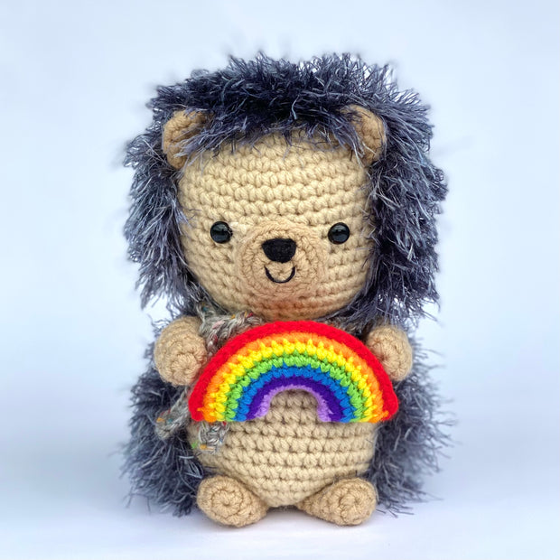 Crocheted plush of a cute hedgehog, sitting up and holding a small rainbow in its hands. It has fluffy exterior fur and a cute smiling face. 