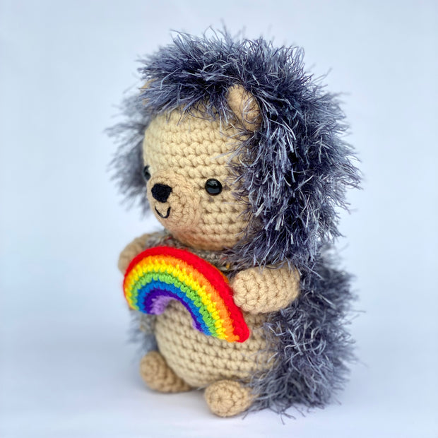 Crocheted plush of a cute hedgehog, sitting up and holding a small rainbow in its hands. It has fluffy exterior fur and a cute smiling face. Side view.