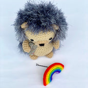 Crocheted plush of a cute hedgehog, sitting up near a small rainbow attached to a bobby pin. It has fluffy exterior fur and a cute smiling face.