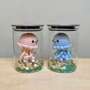 2 glass jars, each with a plush jellyfish inside with a cute face sewn on. At the bottom of each jar are colorful rocks. One jellyfish is pink and the other is blue.