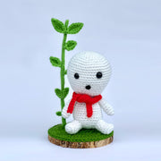 Crocheted sculpture of a small white simplistic forest spirit with a red scarf, sitting on a slice of wood with a large sprout growing from it.