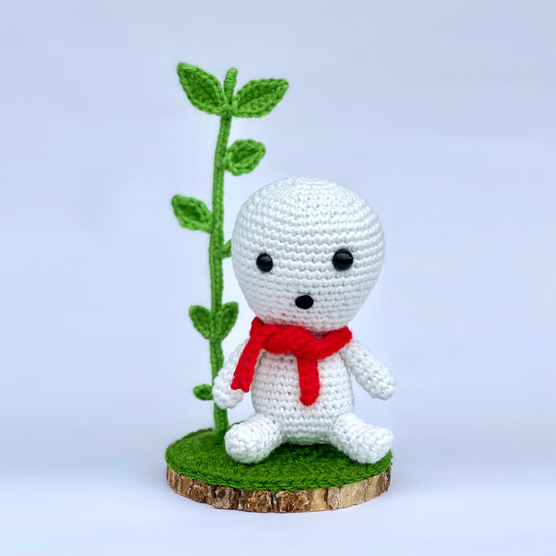 Crocheted sculpture of a small white simplistic forest spirit with a red scarf, sitting on a slice of wood with a large sprout growing from it.
