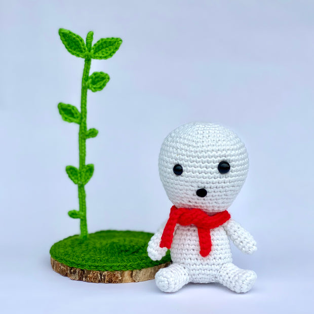 Crocheted sculpture of a small white simplistic forest spirit with a red scarf, sitting next to a slice of wood with a large sprout growing from it.