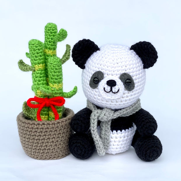 Crocheted sitting panda with a grey scarf and a cute smiling face, sitting next to a potted bamboo plant with many sprouts and a red ribbon tied around it.