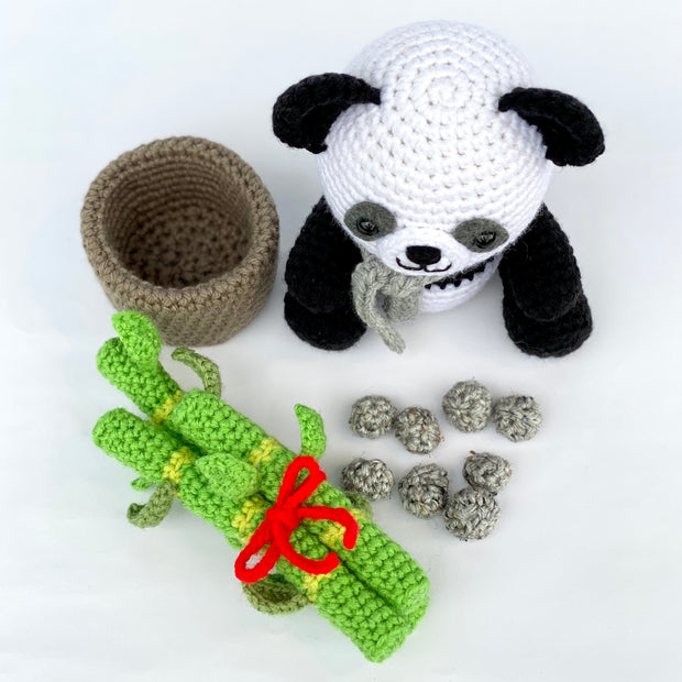 Overhead view of all the components of a crocheted sculpture consisting of a panda, an empty pot, a bundle of sprouting bamboo tied with a red ribbon and several stones to fill the pot.