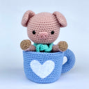 Crochet sculpture of a cute pink pig with an oversized head and a teal scarf around its neck sitting inside of a blue coffee mug. The mug is also crocheted and has a white heart as the center design.