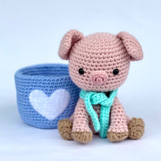  Crochet sculpture of a cute pink pig with an oversized head and a teal scarf around its neck sitting next to a crocheted blue mug with a white heart on it.