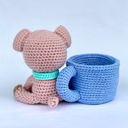  Crochet sculpture of a cute pink pig with an oversized head and a teal scarf around its neck sitting next to a crocheted blue mug with a white heart on it.