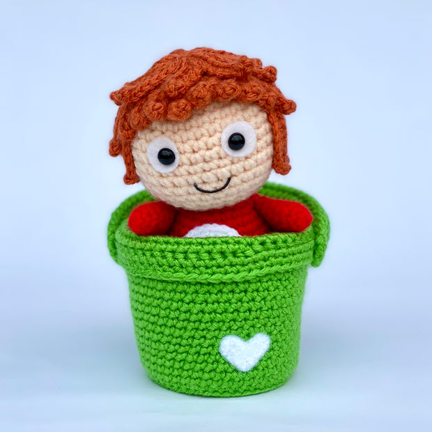 Crochet sculpture of a simply designed redhead girl with a red body, aka Ponyo, sitting in a green crocheted bucket. The bucket has a small white heart in the bottom corner.