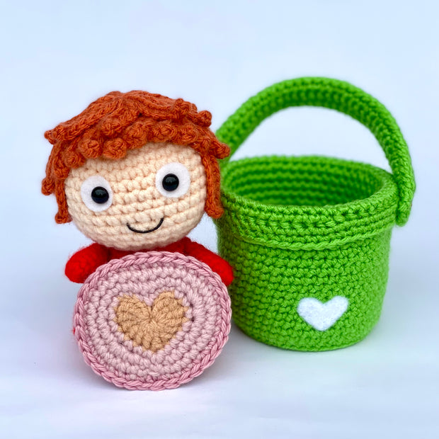 Crochet sculpture of a simply designed redhead girl with a red body, aka Ponyo next to an empty crocheted green bucket. In Ponyo's hands is a pink crocheted coaster with a heart in the center.