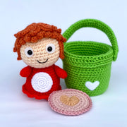 Crochet sculpture of a simply designed redhead girl with a red body, aka Ponyo next to an empty crocheted green bucket. Near Ponyo is a pink crocheted coaster with a heart in the center.