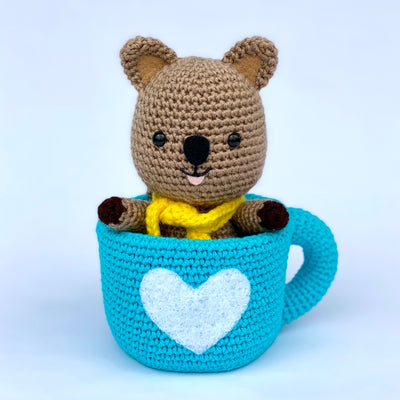 Crochet sculpture of a quokka, a small brown marsupial, wearing a yellow knit scarf and sitting in a blue crocheted mug with a white heart as the center design.