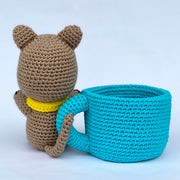 Crochet sculpture of a quokka, a small brown marsupial, wearing a yellow knit scarf and sitting outside of a blue crocheted mug with a white heart as the center design. Back view.