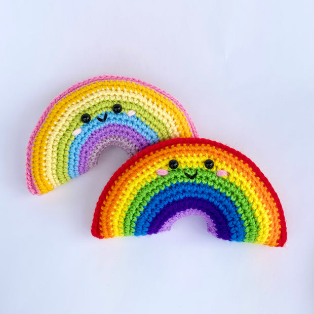 2 crocheted rainbows with simple, cute smiling faces with pink blushing cheeks. One rainbow consists of traditional colors and the other consists of pastel rainbow colors.