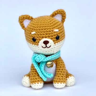 Crocheted shiba inu dog with a slightly oversized head, sitting with a turquoise scarf around its neck and a gold metal bell. It has a cute, simple smiling face.