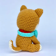 Crocheted shiba inu dog with a slightly oversized head, sitting with a turquoise scarf around its neck and a gold metal bell. Back view.