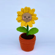 Crochet sunflower, growing out of a crocheted clay plot with 2 leaves growing from the stem. Sunflower has a cute smiling face with pink cheeks.