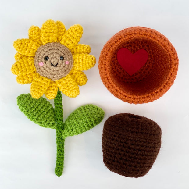 Unassembled crochet sculpture, displaying a smiling sunflower, an empty crocheted pot and a brown mound of soil.