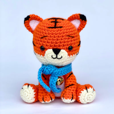 Crocheted tiger with a slightly oversized head and a sweet, smiling expression. It sits and wears a blue knit scarf with a metal bell around its neck.