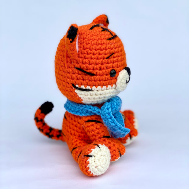 Crocheted tiger with a slightly oversized head and a sweet, smiling expression. It sits and wears a blue knit scarf with a metal bell around its neck. Side view.