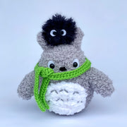 Crochet sculpture of Totoro, with a short rounded body and a green scarf wrapped around his neck, blocking his mouth. Atop his head is a fluffy black dust sprite.