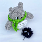 Crochet sculpture of Totoro, with a short rounded body and a green scarf wrapped around his neck, blocking his mouth. Nearby is a fluffy black dust sprite attached to a bobby pin.