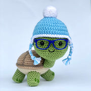 Crochet of a cute, smiling turtle with a slightly oversized head. It stands on 4 feet and has blue glasses. Atop its head is a blue beanie with a white pom pom at the top.