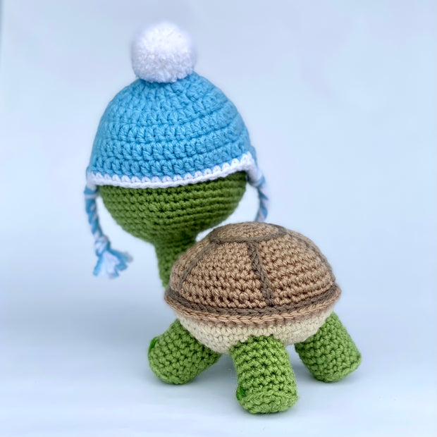 Crochet of a cute, smiling turtle with a slightly oversized head. It stands on 4 feet and has blue glasses. Atop its head is a blue beanie with a white pom pom at the top. Back view.
