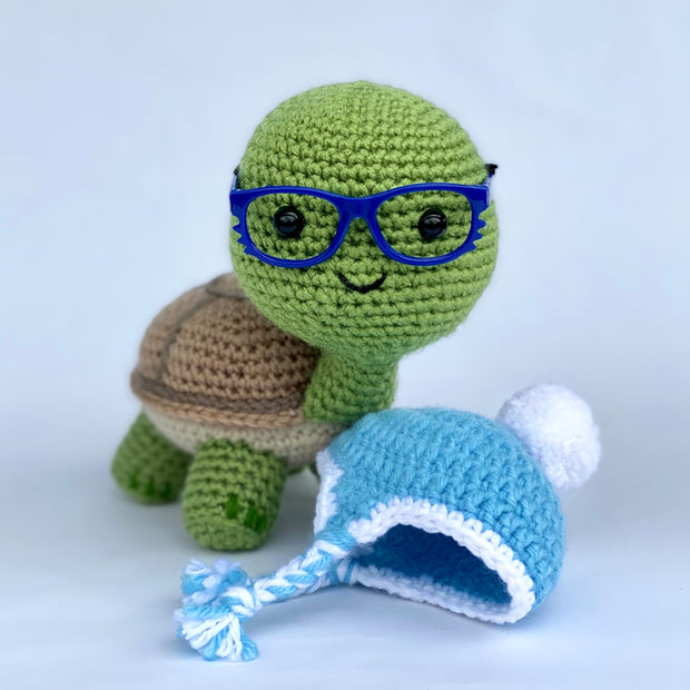 Crochet of a cute, smiling turtle with a slightly oversized head. It stands on 4 feet and has blue glasses. Nearby is a blue beanie with a white pom pom at the top.