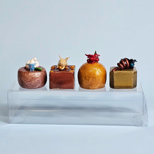 4 mini sculptures of realistic chocolate bon bons, glazed with shiny finishes. Each is adorned by a small winged creature and other thematic candy toppings.