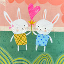 Illustration of 2 cute cartoon style white bunnies, each wearing a patterned dress and smiling. Together, they hold a flower with a large heart ballooning out the middle. They stand in a grassy field with a pink background and green striping pattern along the bottom.