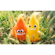 2 very fluffy flame plushies, one orange with a smiling underbite and one yellow with 2 buckteeth. Both have large plastic eyes and sit in a grassy field.