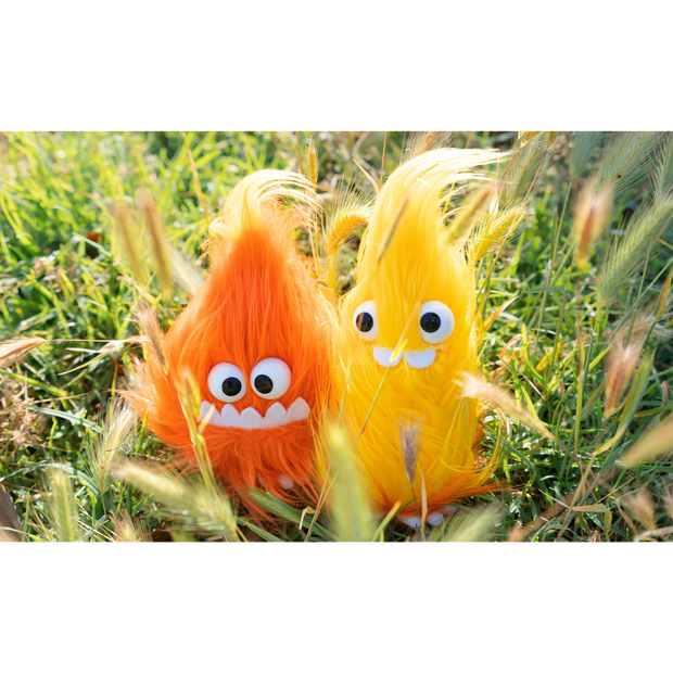 2 very fluffy flame plushies, one orange with a smiling underbite and one yellow with 2 buckteeth. Both have large plastic eyes and sit in a grassy field.