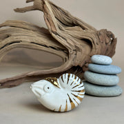 Ceramic figurine of a nautilus with gold stripe detailing. It sits in front of a setting with stacked stones and driftwood.
