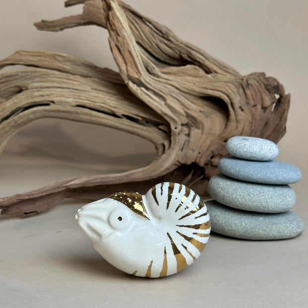 Ceramic figurine of a nautilus with gold stripe detailing. It sits in front of a setting with stacked stones and driftwood.