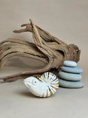 Ceramic figurine of a nautilus with gold stripe detailing. It sits in front of a setting with stacked stones and driftwood.