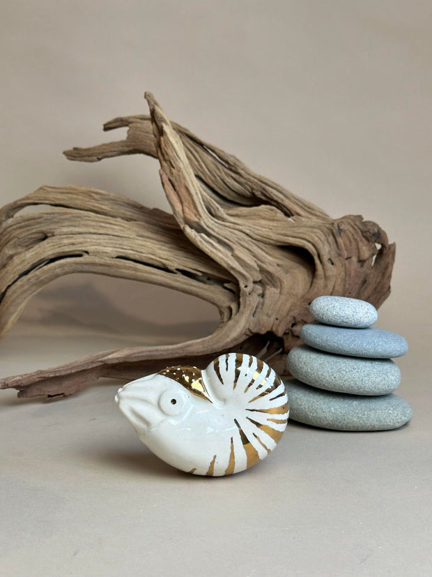 Ceramic figurine of a nautilus with gold stripe detailing. It sits in front of a setting with stacked stones and driftwood.
