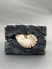 Ceramic figurine of a nautilus with gold stripe detailing. It is encased in a block of black styrofoam that looks like a molten rock.