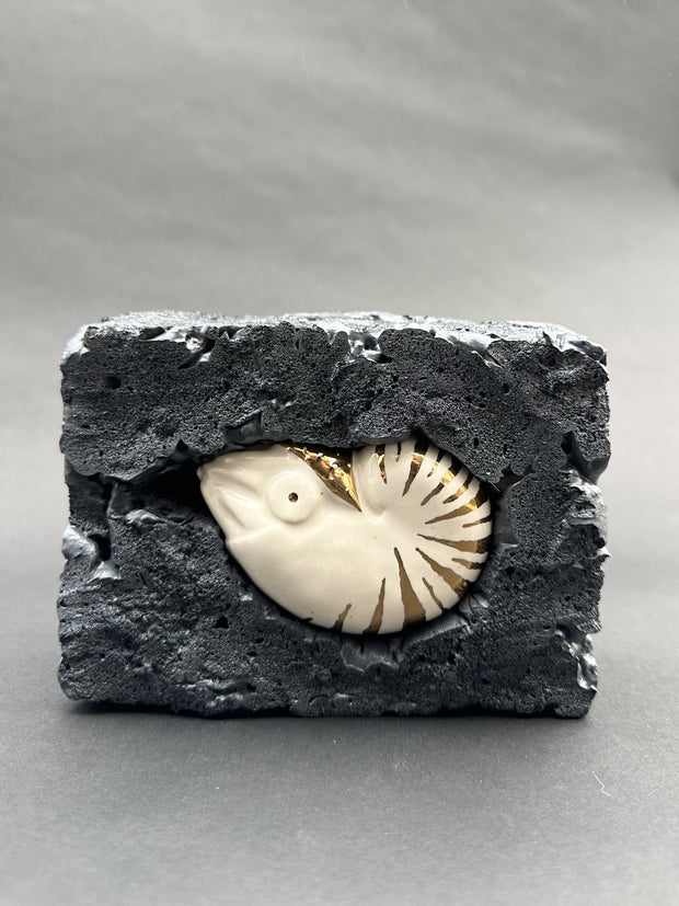Ceramic figurine of a nautilus with gold stripe detailing. It is encased in a block of black styrofoam that looks like a molten rock.