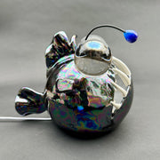 Ceramic sculpture with a pearlescent finish of a round bodied angler fish, with giant bulbous eyes and a blue antenna overhead.