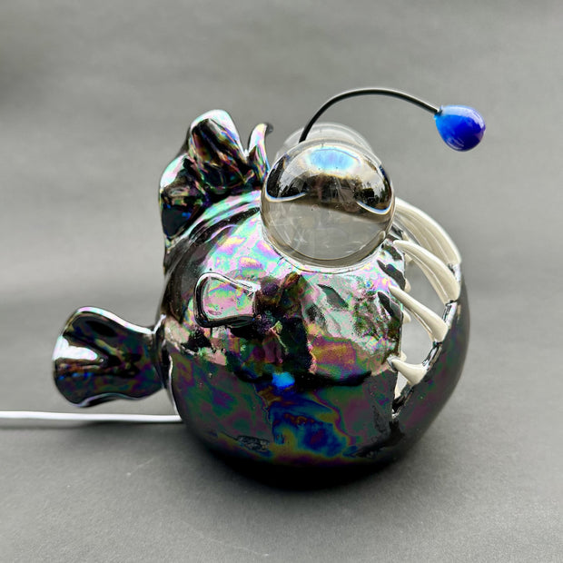 Ceramic sculpture with a pearlescent finish of a round bodied angler fish, with giant bulbous eyes and a blue antenna overhead.