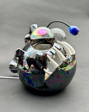 Ceramic sculpture with a pearlescent finish of a round bodied angler fish, with giant bulbous eyes and a blue antenna overhead.