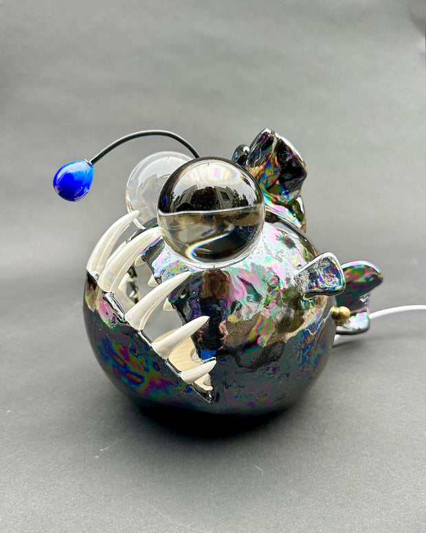 Ceramic sculpture with a pearlescent finish of a round bodied angler fish, with giant bulbous eyes and a blue antenna overhead.
