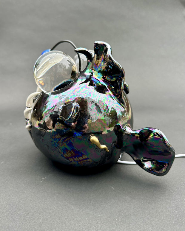 Ceramic sculpture with a pearlescent finish of a round bodied angler fish, with giant bulbous eyes and a blue antenna overhead.