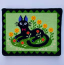 Embroidered artwork of a black cat resting in the flowers on a green background. The cat has a red bowtie.