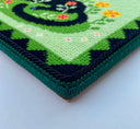 Embroidered artwork of a black cat resting in the flowers on a green background. The cat has a red bowtie. Close up detail of side.
