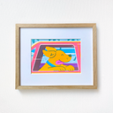 A framed art print featuring a colorful cartoon dog peeking out of a pink car window, mounted on a white wall with a light wood frame and white mat.