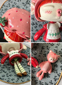 Ceramic sculpture of a doll with a cute, large closed eye head. It is dressed like Strawberry Shortcake and mounted to a wood plaque with a small bear.