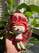 Ceramic sculpture of a doll with a cute, large closed eye head. It is dressed like Strawberry Shortcake and mounted to a wood plaque with a small bear.